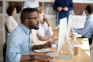 focused african employee wearing glasses looking at computer screen sitting in shared office. serious black manager holding pen make some notes on textbook using pc working on project analyzing data