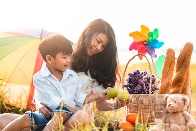 little asian boy sitting on the mother's lap at wide green field when going to picnic. mom and son playing together and looking green apples at hand.celebrating in mother day and appreciating concept.