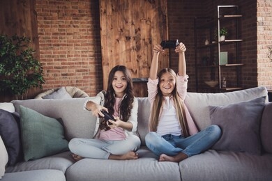 portrait of two nice pretty lovely sweet adorable attractive charming funny cheerful positive girls sitting on divan playing video game battle crossed legs in house loft industrial interior style