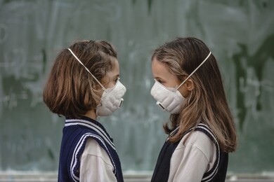 two girls in school uniforms with air pollution masks on face look at each other, standing in front of chalkboard in classroom.