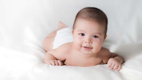 happy cute 5 month old asian baby boy with short black hair wearing a white cloth nappy and lying on his front on a white bed