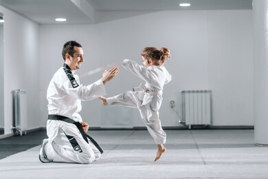 smiling caucasian taekwondo trainer in dobok kneeling and holding hand up while little girl kicking him.
