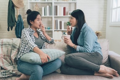 shocked young woman telling her girlfriend some surprised secret. two asian girls gossiping. excited emotional female talking chatting to friend while sitting on couch sofa in living room at home.