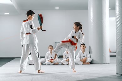 caucasain boy and girl in doboks having taekwondo training at gym. girl kicking while boy holding kick target. in background their friend sitting with legs crossed and watching them.