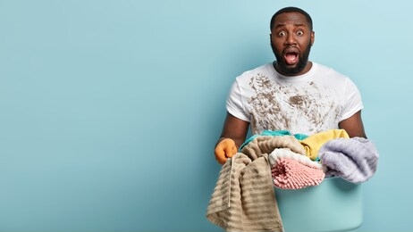 hard working frustrated dark skinned man has dirty white t shirt after cleaning house, holds huge heavy laundry basket with linen, busy doing housework, stares at camera, isolated on blue wall