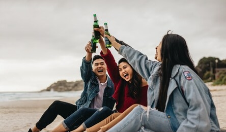 group of four asian friends drinking beer on the sea shore. young people on the beach having a party with drinks.