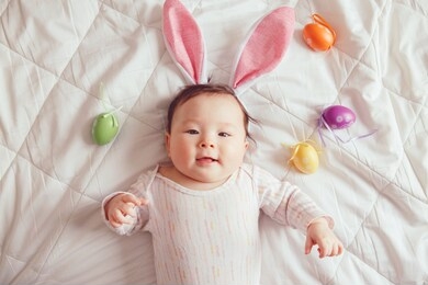 cute adorable mixed asian baby wearing pink easter bunny ears lying on bed in bedroom with colorful eggs. funny kid infant celebrating traditional christian holiday. view from top above