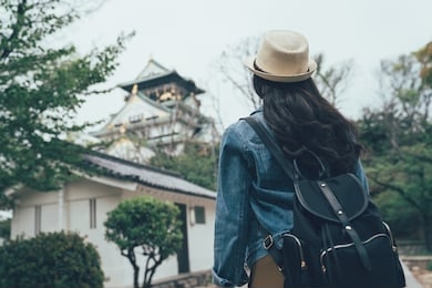 back view of young asian woman traveler wear hat with backpack on shoulder out sightseeing foreign old city in japan. female chinese examines architectural monument osaka castle standing in garden.