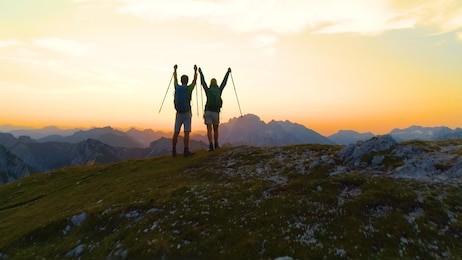 aerial: unrecognizable young hiker couple outstretch arms as they catch the sunset while hiking in the julian alps. picturesque shot of golden sunrise illuminating the excited male and female trekkers