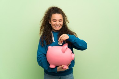 teenager girl over green wall taking a piggy bank and happy because it is full