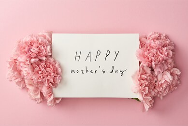 top view of greeting card with happy mothers day lettering and pink carnations on pink background