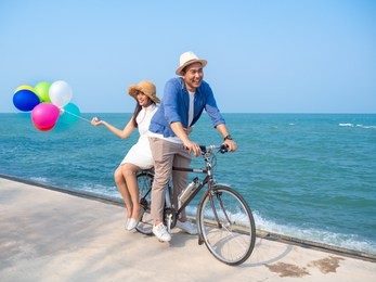 happy asian couple riding bicycle and holding colorful balloons on the beach, lifestyle concept.