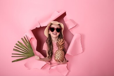 funny little girl peeking out of a hole in a beach hat and heart-shaped glasses with pineapple in her hands and tropical leaves, on a colored background, space for text, studio shooting
