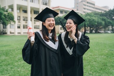 graduate student woman happy talk using smartphone smiling. young college girl friends having phone call laughing cheerfully wearing academic dress standing on lawn. summer graduation season nature