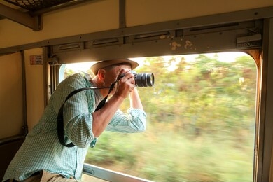 smile asian male traveler with hat has fun trip by train, extending head outside the window for taking pictures by camera.