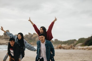 two young couples walking by the beach, with men carrying their women on their back. couples piggybacking on sea shore.