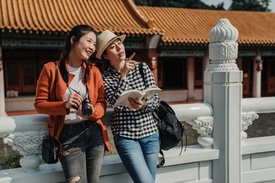 young asian women travelers holding guide book in hands pointing direction to female photographer friend. girls showing place searching destination outdoor discussing leaning on white marble railings