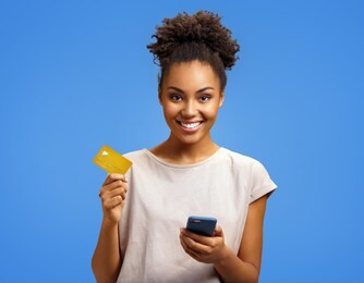 smiling girl holds smart phone and credit card. photo of african american girl wears casual outfit on blue background. emotions and pleasant feelings concept.