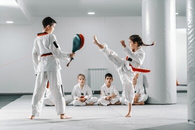 caucasain boy and girl in doboks having taekwondo training at gym. girl kicking while boy holding kick target. in background their friend sitting with legs crossed and watching them.
