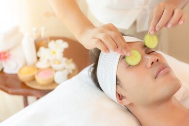 massage therapist using cucumber slices put to handsome young asian man eyes for restore moisture of eyes skin and solve dark circles under eyes. attractive handsome young guy use service at spa salon