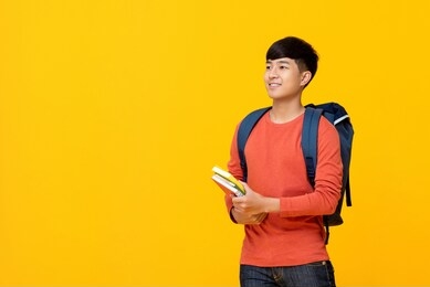 young smiling handsome asian male college student with backpack holding books studio shot for education concept
