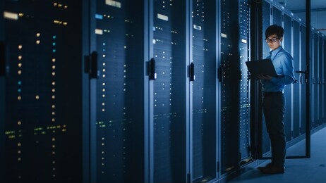 in dark data center: male it specialist stands beside the row of operational server racks, uses laptop for maintenance. concept for cloud computing, artificial intelligence, cybersecurity.