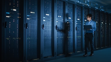 in dark data center: male it specialist walks along the row of operational server racks, uses laptop for maintenance. concept for cloud computing, artificial intelligence, supercomputer, cybersecurity