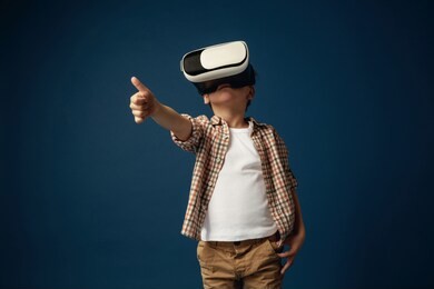 another view of the world. little boy or child in jeans and shirt with virtual reality headset glasses isolated on blue studio background. concept of cutting edge technology, video games, innovation.
