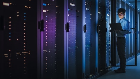in dark data center: male it specialist stands beside the row of operational server racks, uses laptop for maintenance. concept for cloud computing, artificial intelligence, supercomputer. neon lights
