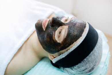 portrait of beautiful woman laying with towel on the head. young girl enjoys black cream facial mask. lady getting spa treatment at beauty salon.