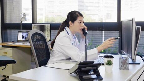 asian woman doctor sitting at desk in front of computer and talking on telephone with a patient telemedicine health hotline consultation. young female medical staff using pen pointing screen discuss