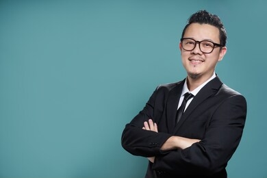 portrait of a smart asian smiling and confident young businessman in black suit and tie keeping arms crossed , looking at camera isolated on vintage blue color background .