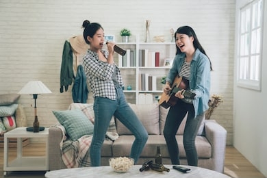 young happy girl best friends having party one playing guitar and another singing by holding beer bottle as microphone. two asian women dancing together. friendship leisure rest home enjoy concept