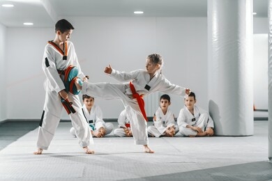 caucasain boy and girl in doboks having taekwondo training at gym. girl kicking while boy holding kick target. in background their friend sitting with legs crossed and watching them.