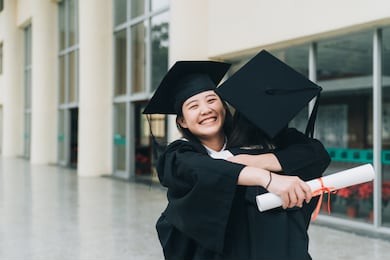 graduates in mantles with diplomas in hands standing near university building and hugging each other. two asian college girl best friends close smiling giving best blessing in graduation ceremony.