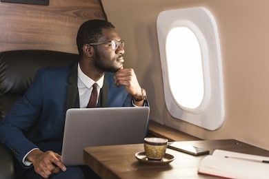 attractive and successful african american businessman with glasses working on a laptop while sitting in the chair of his private jet.