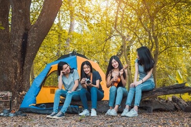 friends group of young asian women camping and resting at forest playing ukulele,take a photo happy on weekend.