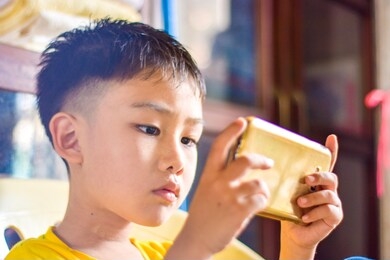 cute asian chinese boy with squint eyes playing mobile game on smartphone that shows eye damage or eye strain