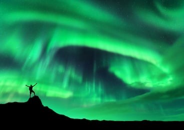 aurora borealis and silhouette of a man with raised up arms on the mountain peak. lofoten islands, norway. aurora and happy man. sky with stars and polar lights. night landscape with aurora and people