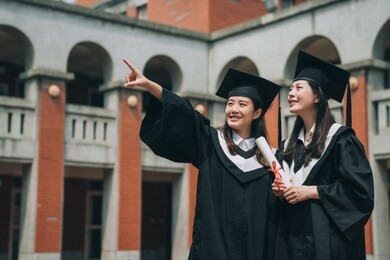 two smiling happy asian women university student standing outdoor in campus with red brick traditional building in back. college girl in gowns showing friends holding diploma point finger at sky.