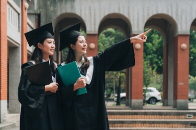 happy asian college girls smiling point finger at sky standing in traditional red brick wall campus outdoor. two university student holding books wear academic dress and mortarboard on graduate.