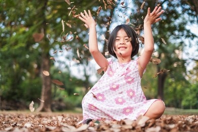 a portrait of adorable girl sitting on grass& throwing tree leaves in forest park. the lovely girl loves outdoor & green nature. children likes having fun in fall. eco friendly& earth day concept.    