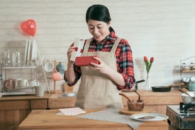 young girl holding box full of chocolate candies prepared valentine gift for boyfriend in kitchen. woman in apron standing near stove smiling looking her handmade sweet cocoa dessert indoor.