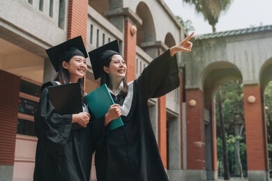 low angle view two asian college girls smiling point finger at sunny blue sky full of hope wish in heart. smart elegant students in graduation gowns and caps holding book stand outdoor red brick wall