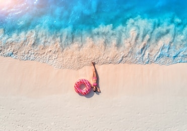 aerial view of the beautiful young lying woman with pink donut swim ring on the white sandy beach near sea with waves at sunset. summer holiday. top view of slim girl, clear azure water. indian ocean
