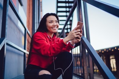 happy japanese hipster girl listening positive audio message from friend during online chatting via smartphone application, cheerful asian millennial enjoying from using modern technology outdoors