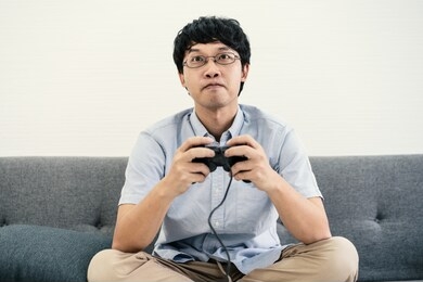 asian man holding joy stick control playing video game on sofa in his room.