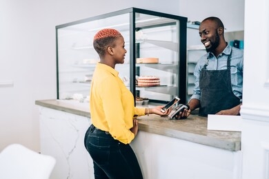 side view of african american female using smartphone to pay for order and looking at smiling male seller while buying food in small restaurant