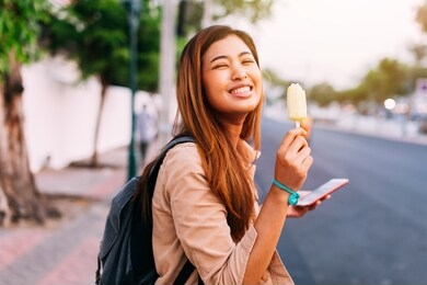 side view of charming asian woman holding smartphone and eating ice-cream while smiling at camera on sunny street