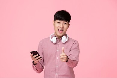 young smiling handsome asian man with headphones and smartphone giving thumbs up studio shot isolated on white background